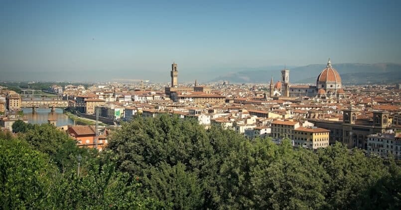Panorama Florencji we Włoszech — widok na katedrę Santa Maria del Fiore z charakterystyczną czerwoną kopułą, dzwonnicę Giotta, Palazzo Vecchio i most Ponte Vecchio nad rzeką Arno, z zielenią w pierwszym planie i górami w tle.