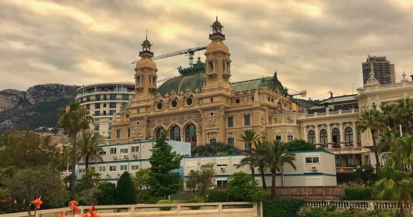 Widok na kasyno Monte Carlo w Monako — historyczny, eklektyczny budynek z zielonym dachem i wieżyczkami, otoczony palmami, ogrodami i nowoczesną zabudową na tle gór i pochmurnego nieba.