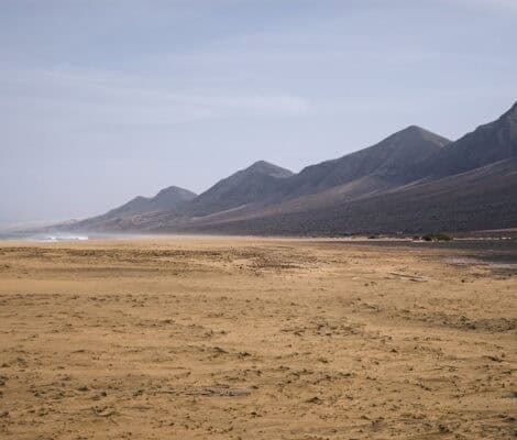 Dzika, rozległa plaża Cofete na Fuerteventurze — złoty piasek, wzburzone fale Atlantyku i surowe góry Jandía w tle, pod zachmurzonym niebem.